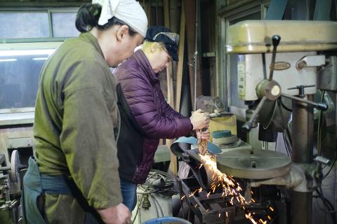 Two individuals are forging a sword in a workshop, with sparks flying from the grinding wheel.