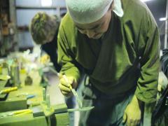 A master swordsmith in a green robe and white headwrap demonstrates a technique on a small sword with a knife, while another person works in the background.