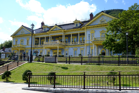 A large, two-story building with blue walls and yellow trim sits on a grassy hill behind a black metal fence under a blue sky with white clouds.