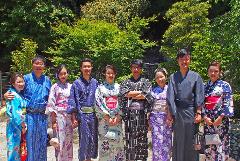 A group of eight people wearing traditional Japanese yukata stand side by side, holding hands and smiling, in front of a lush green background.
