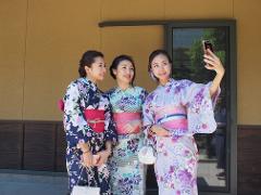 Three smiling Asian women in floral kimonos pose for a selfie in front of a modern building.