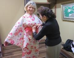 A woman in a floral kimono is being adjusted by another woman in a black shirt and tan pants in a room with wooden floors.
