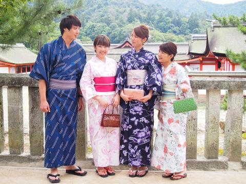 A group of four people wearing kimonos are standing together in front of a stone fence with Japanese writing.