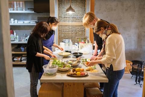 Four people wearing face masks are gathered around a table, preparing food together, with bowls of vegetables and fruit scattered on the table's surface.
