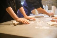 Two people are using rolling pins to flatten dough on a floured surface, likely preparing food in a kitchen or cooking class setting.