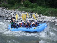 A group of people are joyfully navigating a river in a blue raft, surrounded by splashing water and rocky shores, with green vegetation in the background.