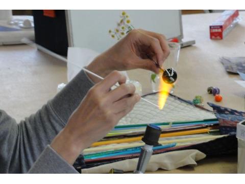 Une personne utilise un chalumeau pour chauffer et façonner une perle de verre, en la tenant avec des tiges au-dessus d&#39;une surface résistante à la chaleur, avec des tiges de verre colorées à proximité.