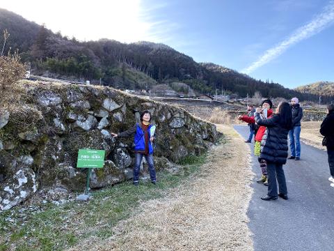 A tour group stands on a path beside a moss-covered stone wall in a rural Japanese village, with a woman pointing at the wall.