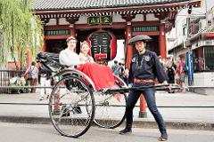 A smiling couple enjoys a rickshaw ride in front of a temple with a rickshaw driver in traditional attire.