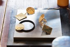 Gold-colored geometric jewelry, including a ring, pendant, and hair tie embellishment, rests on a metal surface next to a wooden table.