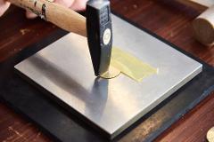 A hammer strikes a metal disc on a steel surface, with masking tape nearby.