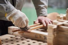 A craftsman's gloved hands use a tool to shape a piece of wood in a workshop, surrounded by wooden components.