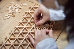 A person's hands assemble a geometric wooden structure from small rectangular pieces.