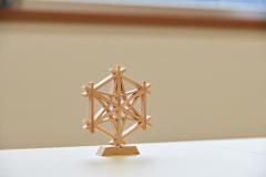 A wooden geometric sculpture is displayed on a light-colored surface with a blurred background.