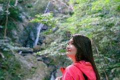 An Asian woman in a pink jacket gazes upwards, standing in front of a lush green forest with a waterfall in the background.
