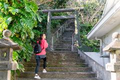A woman with a backpack is walking up stone steps towards a torii gate, surrounded by lush greenery and stone lanterns.