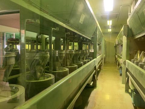 Nishio Matcha Making & Tea Field Walk – Aichi Cultural Tour A row of industrial machinery, likely for processing matcha, is visible through a glass partition, illuminated by overhead lights.