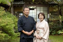 Discover the Art of Yamanaka Lacquerware & Experience the Ultimate Hospitality of the Tea Ceremony An older couple in traditional Japanese attire stands smiling in front of a traditional Japanese building.