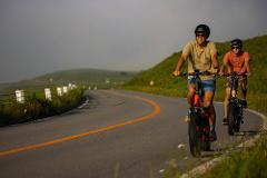 Two people are riding e-bikes on a curved road next to a grassy hillside under a misty sky.