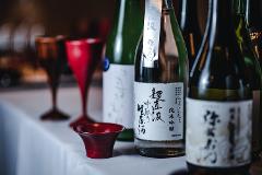 Savor the Essence of Kitakata: A 2-day Luxurious Sake and Lacquerware Experience A close-up shot of several bottles of sake, one with a silver label featuring Japanese characters, sits on a white tablecloth with a red sake cup in the foreground.