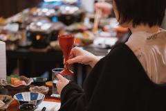 Savor the Essence of Kitakata: A 2-day Luxurious Sake and Lacquerware Experience A woman in traditional attire holds a red sake cup, seated at a table laden with various Japanese dishes and lacquerware.