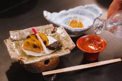 Savor the Essence of Kitakata: A 2-day Luxurious Sake and Lacquerware Experience A hand pours sake from a glass decanter into a small red cup, while a plate of food sits to the left.