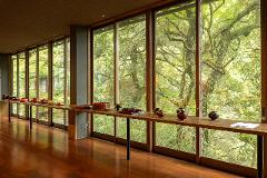 Discover the Art of Yamanaka Lacquerware & Experience the Ultimate Hospitality of the Tea Ceremony A long wooden table displays various lacquerware items in front of a wall of large windows overlooking a lush green forest.