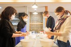 Four masked people are preparing food in a kitchen, pouring ingredients into large white bowls on a light-colored countertop.