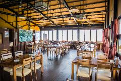 An interior view of a restaurant featuring wooden tables and chairs, large windows offering a view, and a high ceiling with exposed lighting and ductwork.