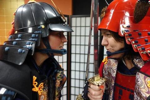 Two people dressed in traditional samurai armor face each other with a sword between them.
