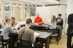 A group of people are gathered around tables in a room decorated for a holiday, some of them are holding snacks.