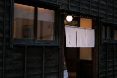 A traditional Japanese storefront with a white noren curtain hangs above an open entrance, illuminated by a soft light, hinting at a culinary establishment.