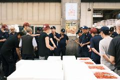 A group of men in a market inspect boxes of shrimp, likely at an auction, with posters in Japanese on a pillar in the background.