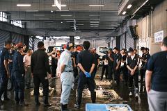 A group of men in uniforms gather around fish displayed in crates at an auction, possibly in a marketplace.