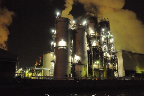 An industrial plant stands illuminated at night, emitting smoke and vapor under a dark sky.
