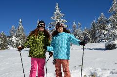 Two women in ski gear pose in the snow with ski poles, surrounded by snow-covered trees under a clear blue sky.