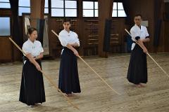 Three individuals in traditional Japanese attire are practicing with bokken swords in a dojo.