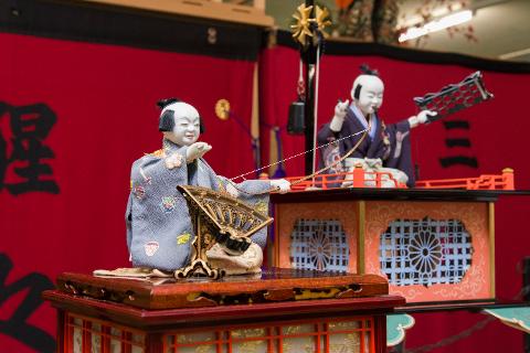 Two Japanese dolls, likely part of a festival display, are adorned in traditional attire and posed with miniature props against a red backdrop.