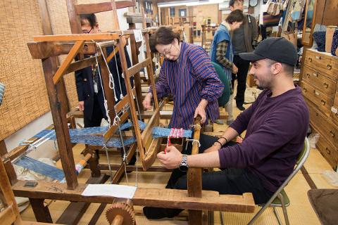 A woman in a striped robe operates a traditional wooden loom as a man in a purple shirt watches.