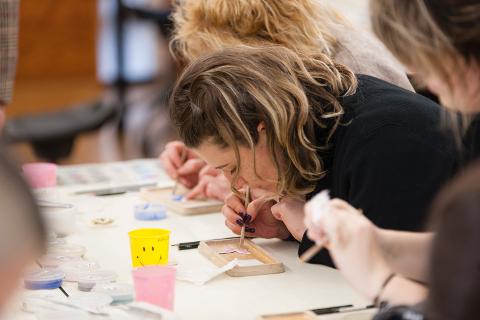 A woman with blonde hair is concentrating on decorating a small metal piece with a stick in an art class.