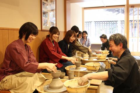 Un gruppo di persone sorridenti è seduto al tornio del vasaio e impara a lavorare la ceramica da un istruttore, anche lui sorridente.
