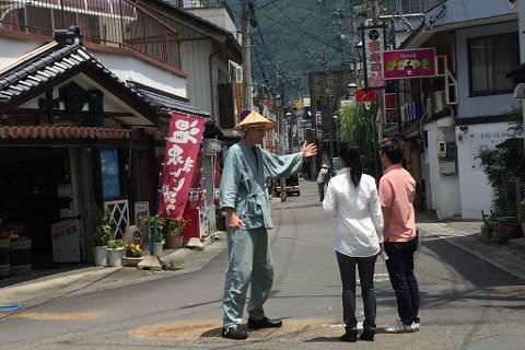 A man in traditional Japanese clothing and a hat is giving directions to two tourists on a narrow street lined with shops and restaurants in a Japanese town.