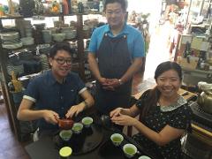 Two people are seated at a table pouring green tea, while a standing person looks on, possibly in a tea shop or similar setting.