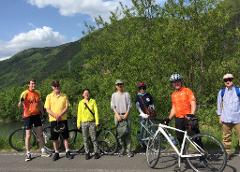 A group of seven people with their bikes pose for a picture on a paved road with a scenic backdrop of trees and mountains.