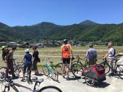 A group of cyclists pause on a concrete path, their bikes lined up as they admire the view of a village, rice paddies, and mountains under a clear blue sky.