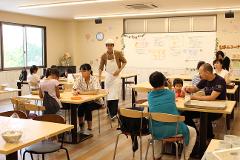 A bright classroom filled with people participating in a baking workshop, led by an instructor in a chef's hat and apron.