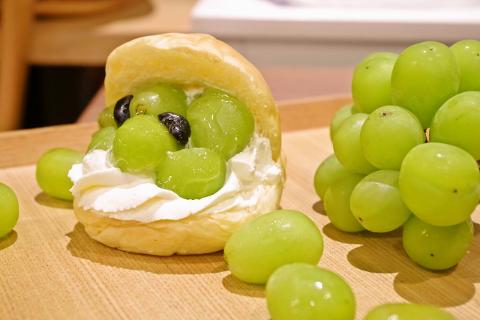 Un gros plan montre un sandwich au pain de couleur claire rempli de crème blanche et de raisins verts avec quelques raisins noirs, à côté d&#39;un tas de raisins verts, sur une surface en bois.
