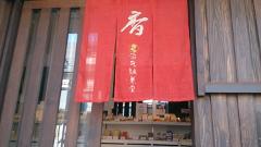 The image shows a traditional Japanese shop entrance with a red noren curtain featuring white calligraphy and a circular emblem, hanging in front of a wooden-framed doorway.