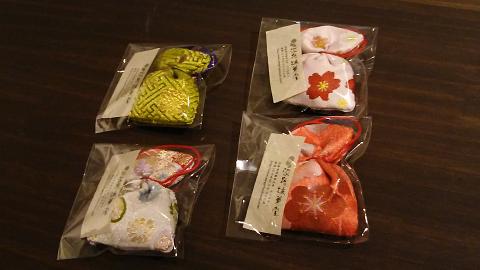 Four wrapped traditional Japanese pouches, in yellow, white, peach, and orange with floral prints, are arranged on a dark wooden surface.