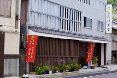 A charming Japanese building with two red banners featuring Japanese text stands on a street lined with potted plants.